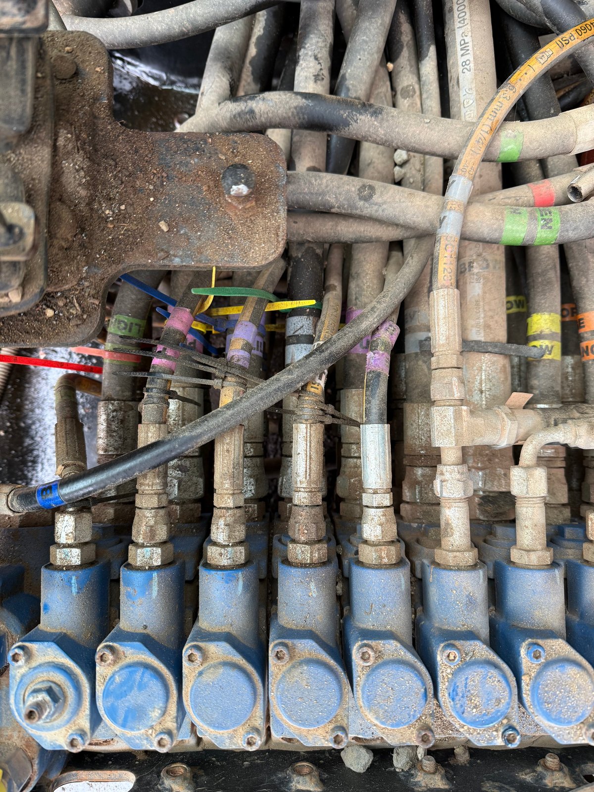 Close-up of hydraulic system components with metal pipes, color-coded hoses, and blue connectors on industrial machinery