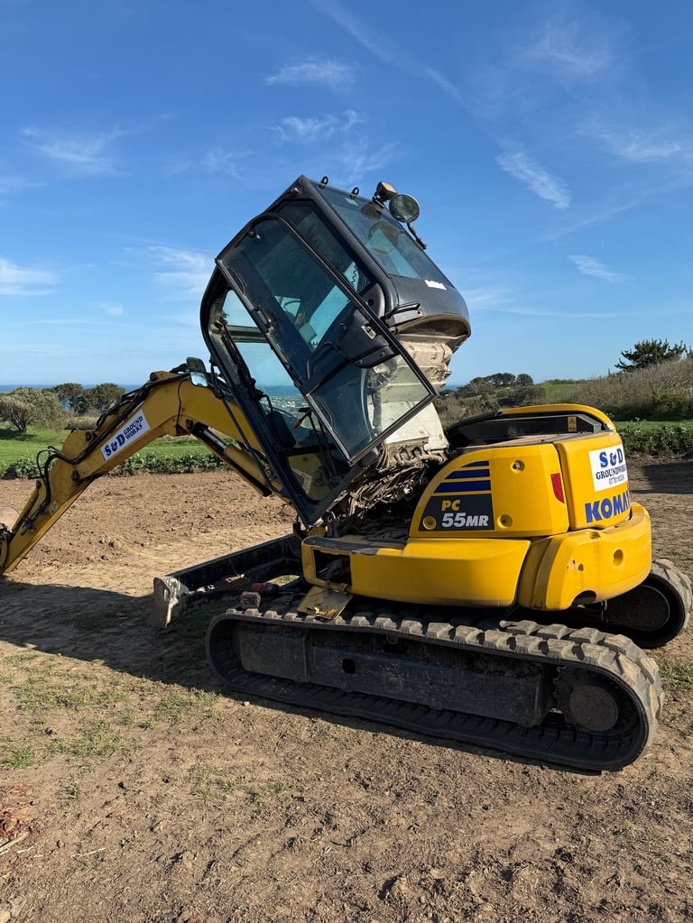 Yellow Komatsu mini excavator with raised arm holding black truck bucket on dirt ground with green landscape and blue sky