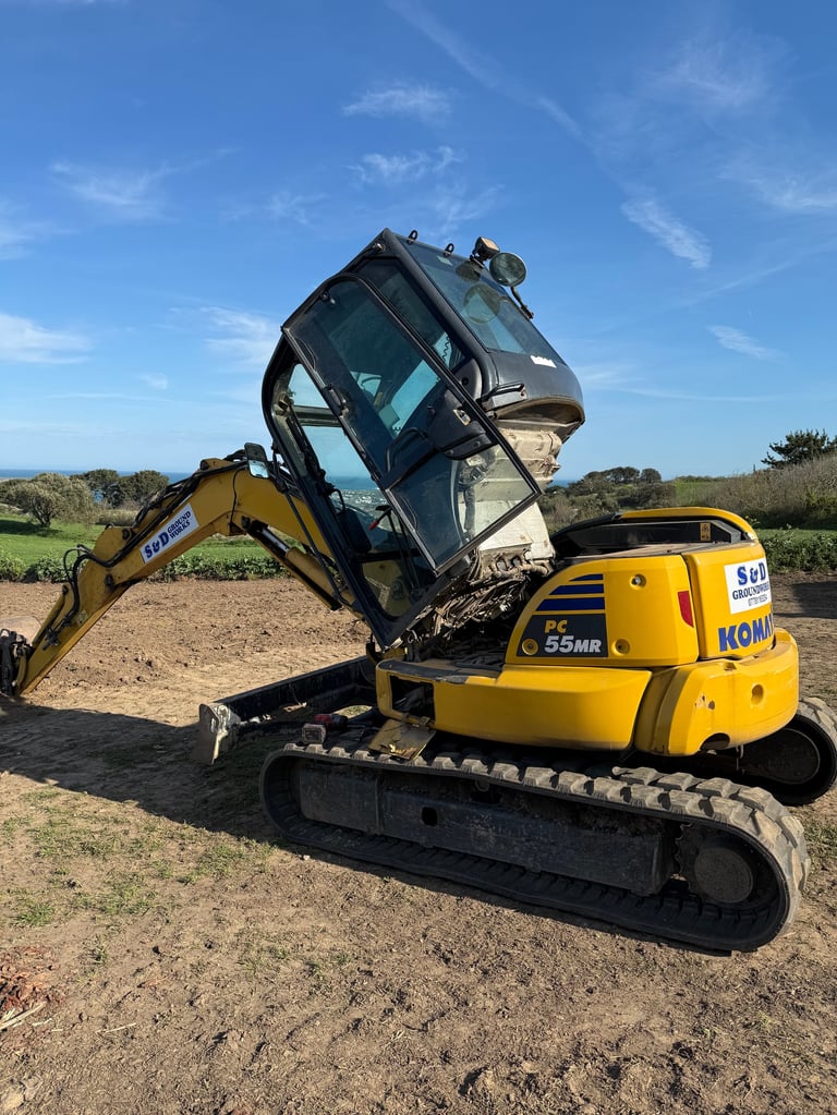 Yellow Komatsu mini excavator lifting a dark-colored car with its articulating arm on a dirt site